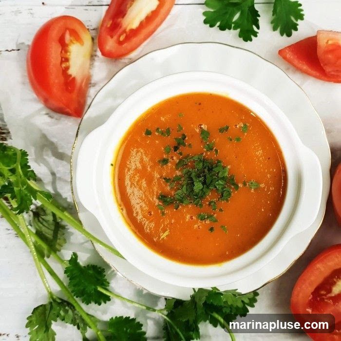 A bowl of creamy tomato soup garnished with chopped parsley, served on a white plate with a gold rim. Sliced tomatoes and cilantro sprigs are arranged around the bowl on a white wooden surface.