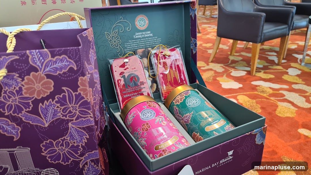 A gift box filled with two decorative tins, individually wrapped cookies, and gift tags. The box and accompanying bag are adorned with a purple batik pattern featuring flowers and Singapore's skyline. The lid of the box has text that reads "SAVOUR THE ICONIC FLAVOURS OF SINGAPORE" and mentions Marina Bay Sands and OSC. One tin is labeled "HAE BEAM" and the other has text that is partially obscured. The bottom of the box displays the "MARINA BAY Sands SINGAPORE" logo.