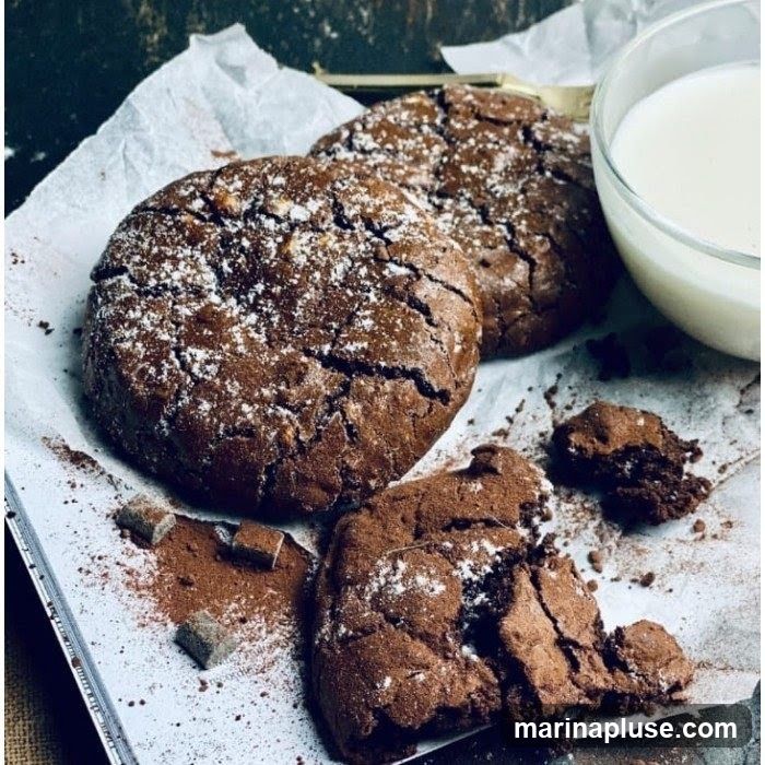 A close-up shot of several chocolate cookies dusted with powdered sugar, arranged on parchment paper. Some cookies are whole, while one is broken, revealing its rich interior. Cocoa powder and sugar cubes are scattered around the cookies. A glass of milk is visible in the upper right corner.