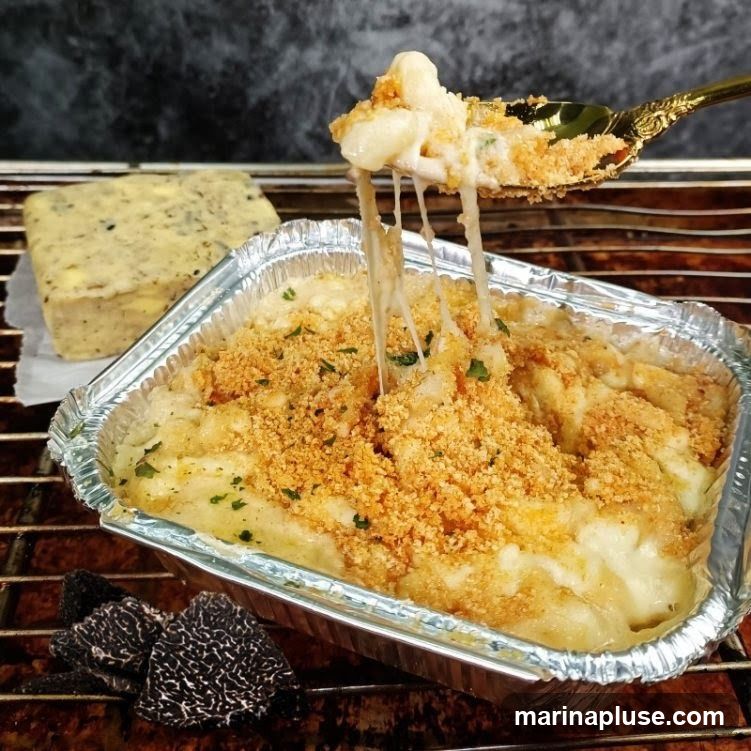 A close-up shot of macaroni and cheese in a foil container, topped with breadcrumbs and parsley. A spoonful of the dish is being lifted, showing melted cheese strings. A block of cheese and truffles are visible in the background.