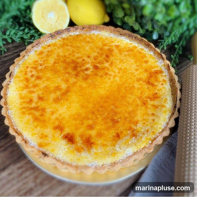 A close-up overhead view of a whole lemon tart with a golden-brown, caramelized sugar topping. The tart has a fluted crust and is garnished with fresh lemons and greenery in the background.