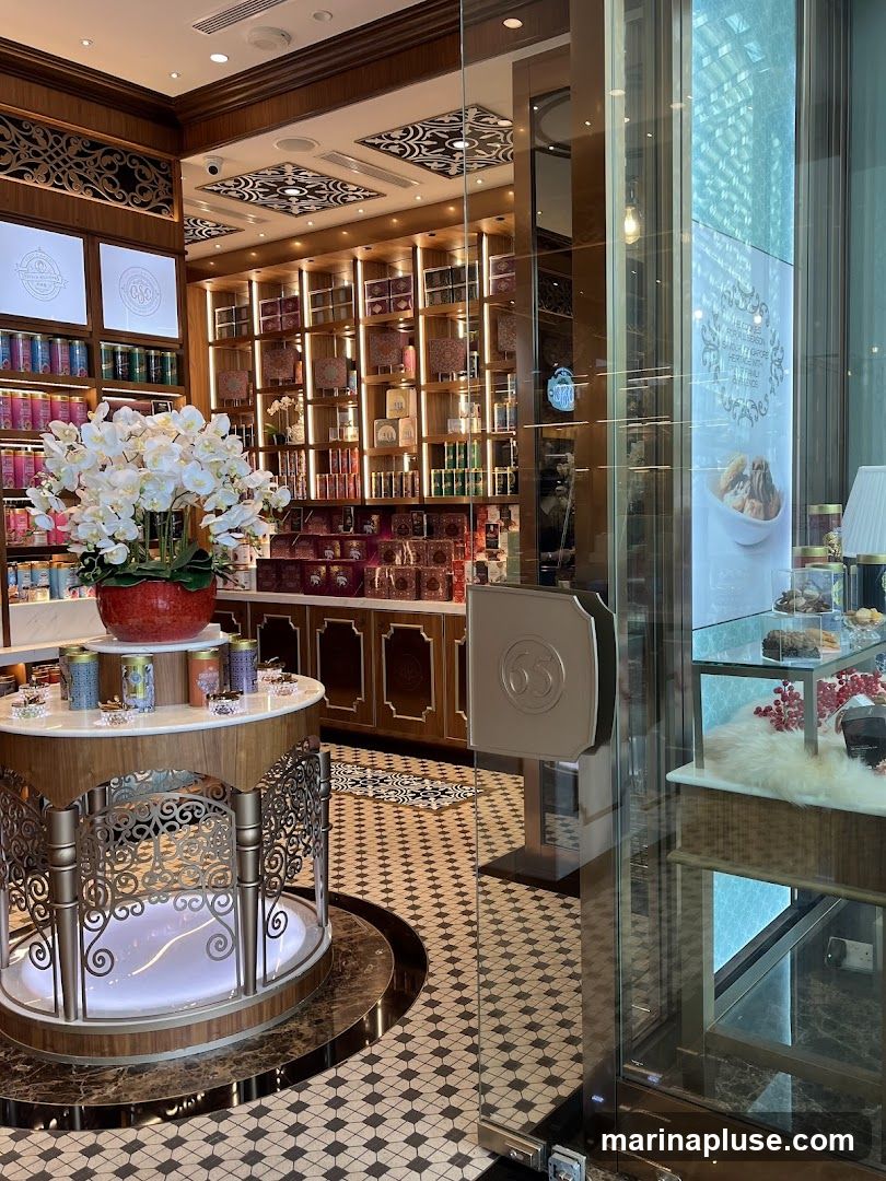 The image shows the interior of a well-lit, opulent confectionery shop. Shelves are stocked with numerous boxes and tins of sweets, likely chocolates or dates, in various sizes and decorative packaging. A central display features a large arrangement of white artificial flowers in a red pot, surrounded by smaller tins. The floor has a distinctive black and white tiled pattern. A glass door with a prominent '65' emblem is visible, suggesting this is an entrance. Through the glass, a display case with more sweets and decorative items can be seen. The overall aesthetic is one of luxury and abundance.