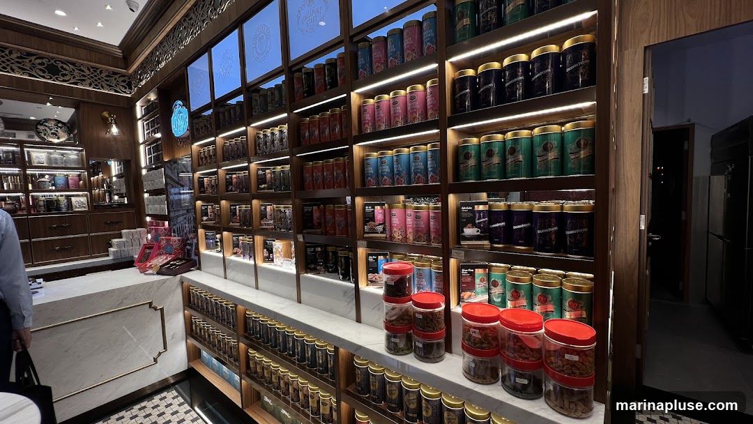 A well-lit interior of a shop, likely a tea or coffee establishment, with extensive shelving filled with numerous cylindrical tins and jars. The shelves are organized and illuminated by strip lighting. A marble counter is visible in the foreground, with a person partially visible on the left. The overall ambiance suggests a premium retail environment focused on beverages and related products.