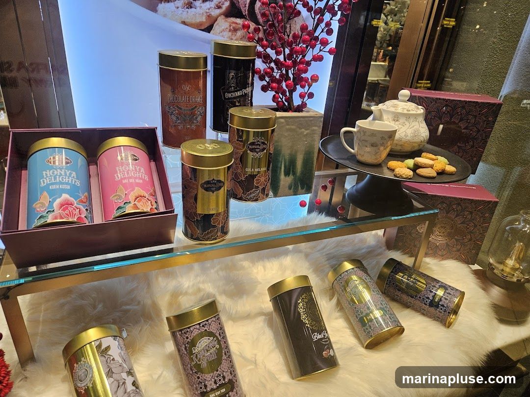 A display of decorative tins filled with tea, along with cookies and a teapot set. The tins feature intricate designs and various flavors like 'NONYA DELIGHTS KUEH KOSUI', 'NONYA DELIGHTS HAE REE HIAM', 'CHOCOLATE ORANGE', and 'ORCHARD PIC'. Some tins are arranged on a white furry surface, while others are on a glass shelf. A teapot, a mug, and a plate of cookies are also visible.