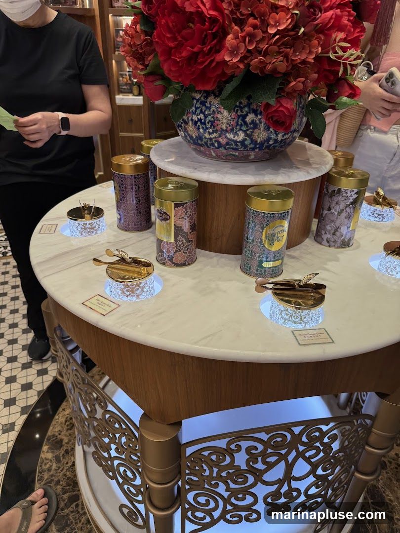 A round marble-topped display table showcases several decorative tins, likely containing tea. A large floral arrangement with red flowers sits atop the display. People are visible in the background, suggesting a retail environment.