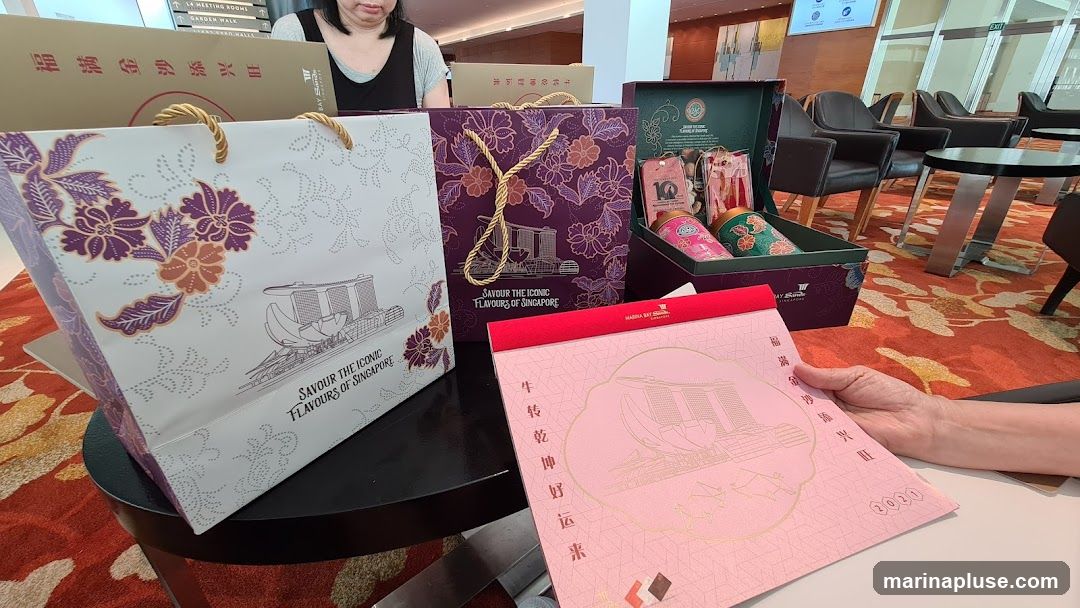 Several gift bags and a gift box are displayed on a table. The bags and box feature designs inspired by Singapore, including the Marina Bay Sands building and floral patterns. One bag has the text "SAVOUR THE ICONIC FLAVOURS OF SINGAPORE". A pink booklet with similar designs and Chinese text is also visible.
