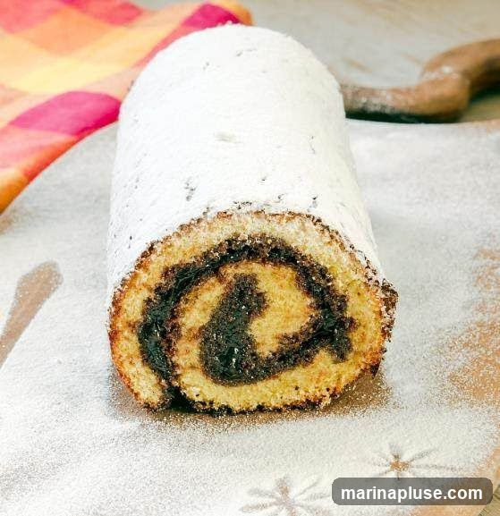 A close-up shot of a Swiss roll cake, generously dusted with powdered sugar. The cake has a light sponge exterior and a dark, swirled filling, likely jam or chocolate. It is presented on a wooden board, with a colorful cloth and a wooden utensil visible in the background.