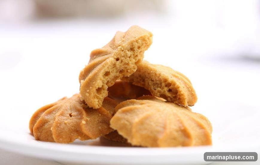A close-up shot of several golden-brown butter cookies stacked on a white plate. One cookie is broken in half, revealing its crumbly texture. The cookies have a ridged, star-like shape. The background is blurred and white.