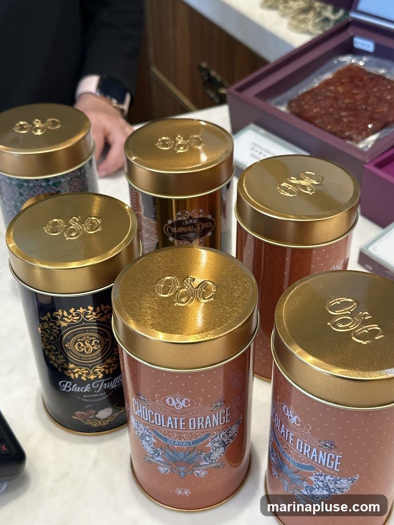 A collection of decorative tins with gold lids, each featuring the embossed logo 'OSC'. One tin is labeled 'Black Truffle with Walnut OSC Cookies'. Two tins are labeled 'Chocolate Orange Seasalt'. Another tin has 'Colonial Days' printed on it. In the background, a person's hand is visible, and a package of dried meat is in a purple box.