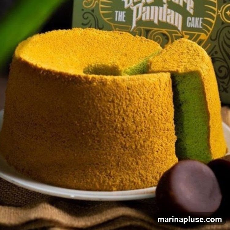A close-up shot of a pandan chiffon cake with a slice removed, revealing its green interior. The cake has a golden-brown exterior and is presented on a white plate. A decorative box with the text "The Pandan Cake" is visible in the background.