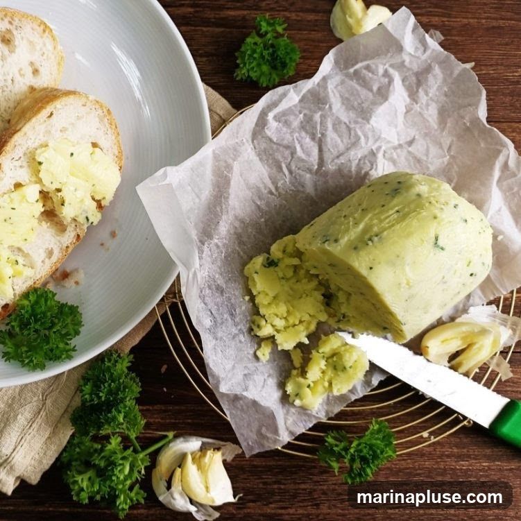 A log of herb butter is presented on parchment paper, with a portion already spread on slices of baguette. Garlic cloves and parsley sprigs are scattered around the serving.