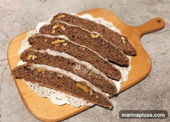 Several slices of dark bread, likely whole wheat or rye, are arranged diagonally on a wooden cutting board. The bread slices are dusted with powdered sugar and contain visible pieces of walnuts. A white, lacy paper doily is placed beneath the bread slices on the board. The background is a textured gray surface.