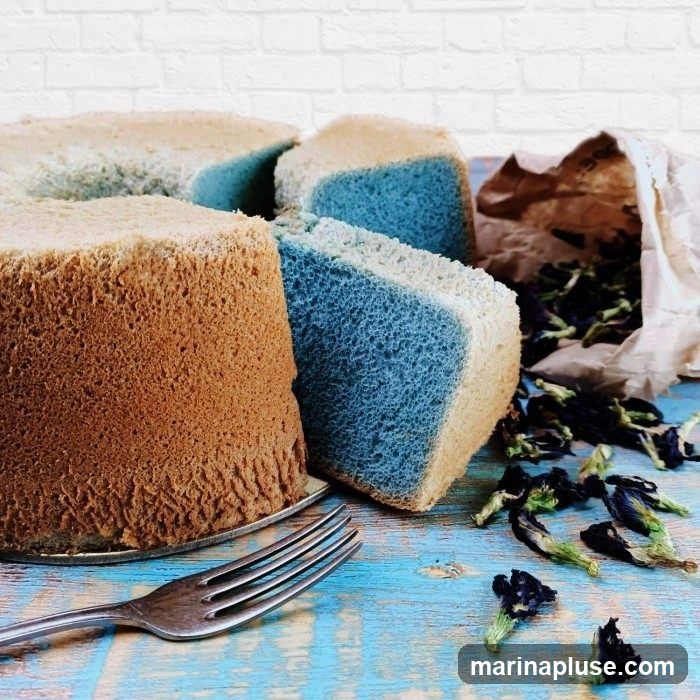 A close-up shot of a chiffon cake that has been cut into slices. Some slices are a light brown color, while others have a blue layer. Dried butterfly pea flowers are scattered on a blue and white wooden surface, with a paper bag in the background. A fork rests in the foreground.