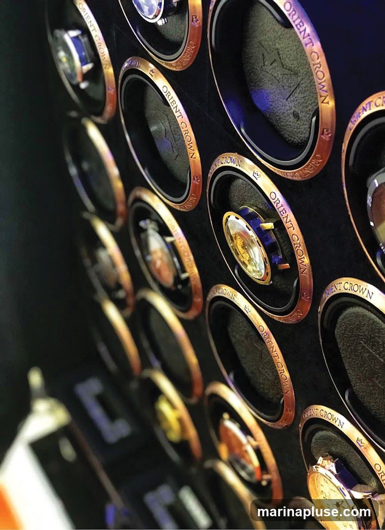 A close-up, angled view of multiple watch winders, each with a copper-colored rim and the text "ORIENT CROWN" inscribed on it. Some winders contain watches, while others are empty. The background is dark and plush.