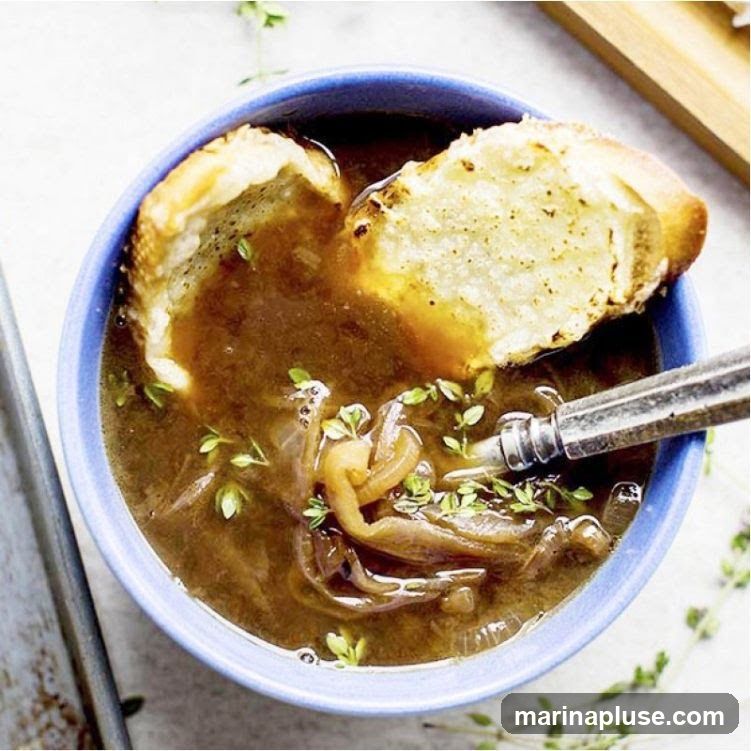 A close-up overhead shot of a bowl of French onion soup. The soup is a rich brown color and filled with thinly sliced onions. Two pieces of toasted bread, likely croutons, are floating on top, one partially submerged. Fresh thyme sprigs are scattered throughout the soup. A silver spoon rests in the bowl, ready to be used. The bowl is a vibrant blue.