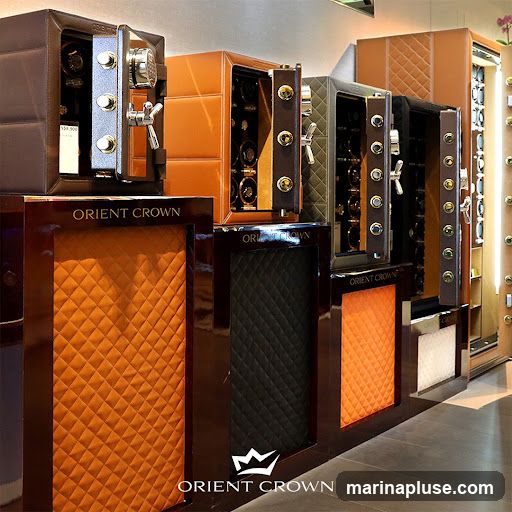 A display of several luxury watch winders, also known as safes, in various colors including brown, tan, dark green, and black. The watch winders are open, revealing compartments designed to hold and wind watches. The brand name 'ORIENT CROWN' is visible on the display stands in front of the watch winders, along with a crown logo. The stands are finished in a dark, glossy wood and feature quilted panels in orange, black, and white.