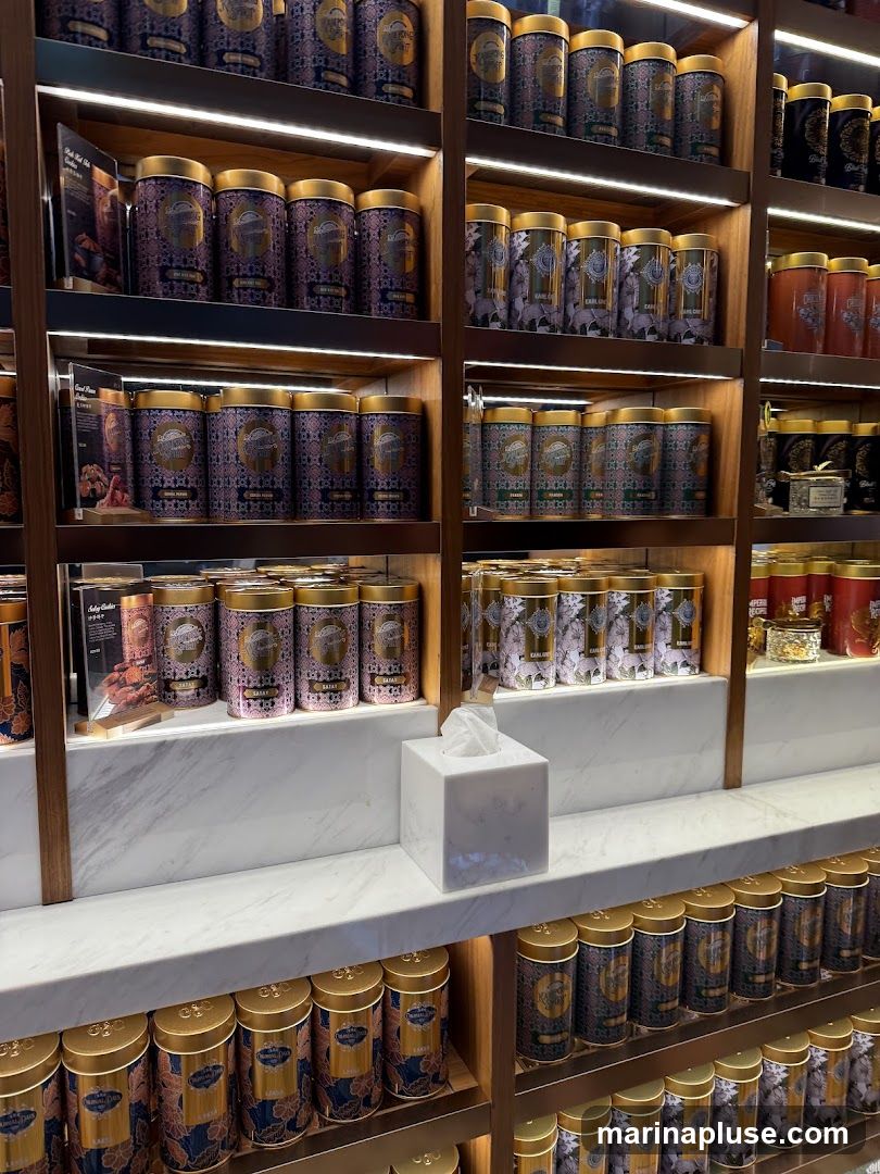 A display of numerous decorative tins, likely containing tea, arranged on wooden shelves with integrated lighting. The tins are of various sizes and designs, featuring intricate patterns in shades of blue, pink, and gold. Some tins have visible labels with text such as 'EARL GREY' and 'SARAY'. A white tissue box sits on a marble shelf in the foreground.