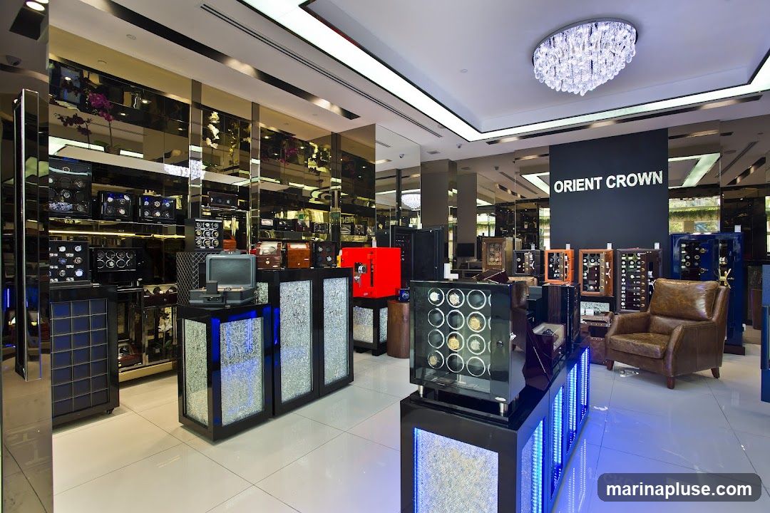 A well-lit interior of a luxury watch store, featuring numerous watch winders and display cases. The walls are lined with mirrored panels, reflecting the merchandise and the opulent crystal chandelier hanging from the ceiling. Prominently displayed on a black panel is the text "ORIENT CROWN". In the foreground, a display case with multiple watch winders is illuminated with blue LED lights. To the right, a plush leather armchair offers a seating area. The store appears to be showcasing a wide selection of high-end timepieces and accessories.