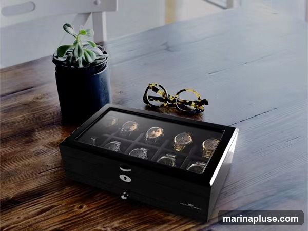 A black wooden watch box with a glass lid sits on a wooden table. Inside the box, twelve watches are displayed on individual cushions. To the right of the watch box, a pair of tortoiseshell glasses rests on the table. To the left, a small potted plant in a black container is visible.