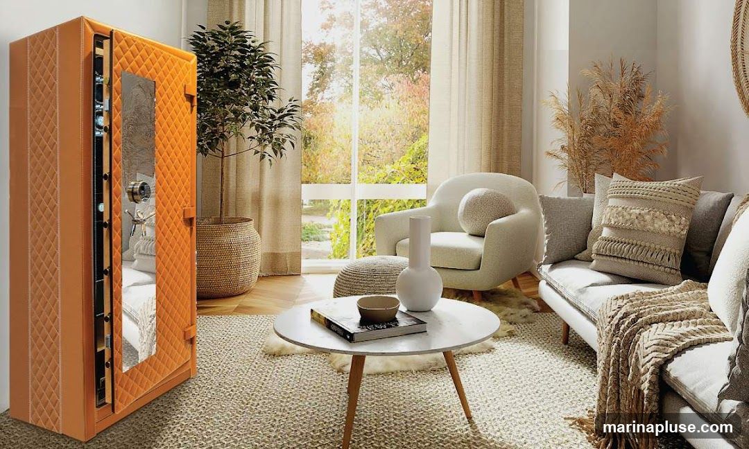 A stylish living room features a large, quilted orange safe with a mirrored door on the left. Inside the safe, several watches are visible. The room is furnished with a comfortable grey sofa adorned with textured cushions and a knitted throw, a cream-colored armchair with a round cushion, and a small round coffee table holding a book and a bowl. A large potted plant stands beside the safe, and dried grasses add a decorative touch. A large window with autumn foliage visible outside provides natural light.