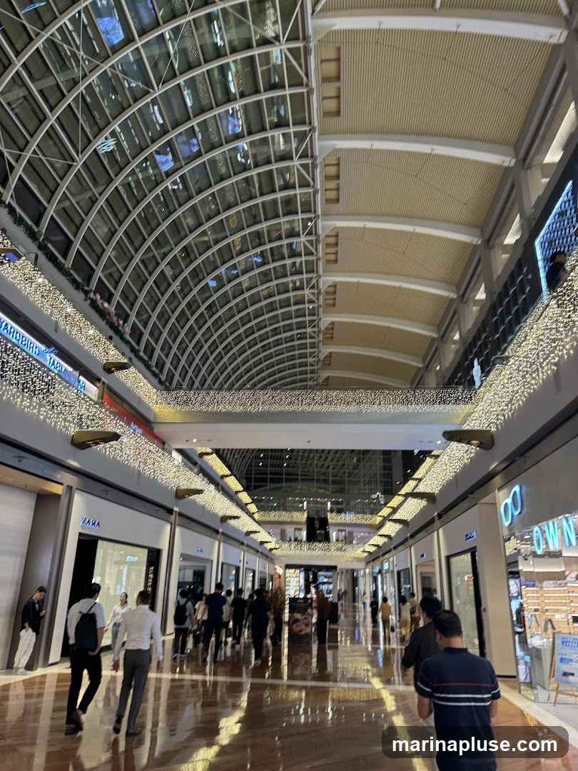 A wide-angle view of a modern shopping mall interior, featuring a high, arched glass ceiling and a long, polished floor reflecting the ambient light. The walkway is lined with various retail stores, some with visible signage like 'ZARA' and 'OWN'. Decorative strings of warm white lights adorn the upper levels of the mall, creating a festive and inviting atmosphere. Several shoppers are seen walking along the corridor, browsing the stores.