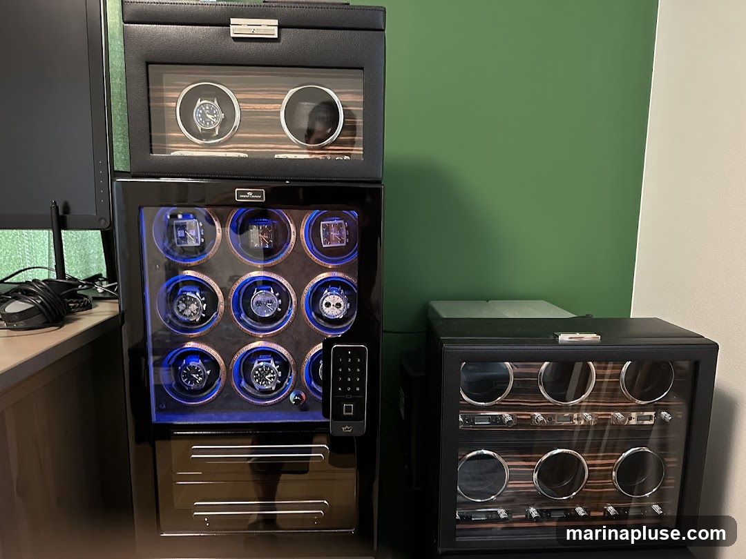 Two black watch winders are shown. The larger one on the left has a top compartment with two watch slots, and a main compartment with nine watch slots, all illuminated with blue light. Below the watch slots are two drawers. A remote control is placed to the right of the main compartment. The smaller watch winder on the right has six watch slots arranged in two rows of three, with wood-grain accents.
