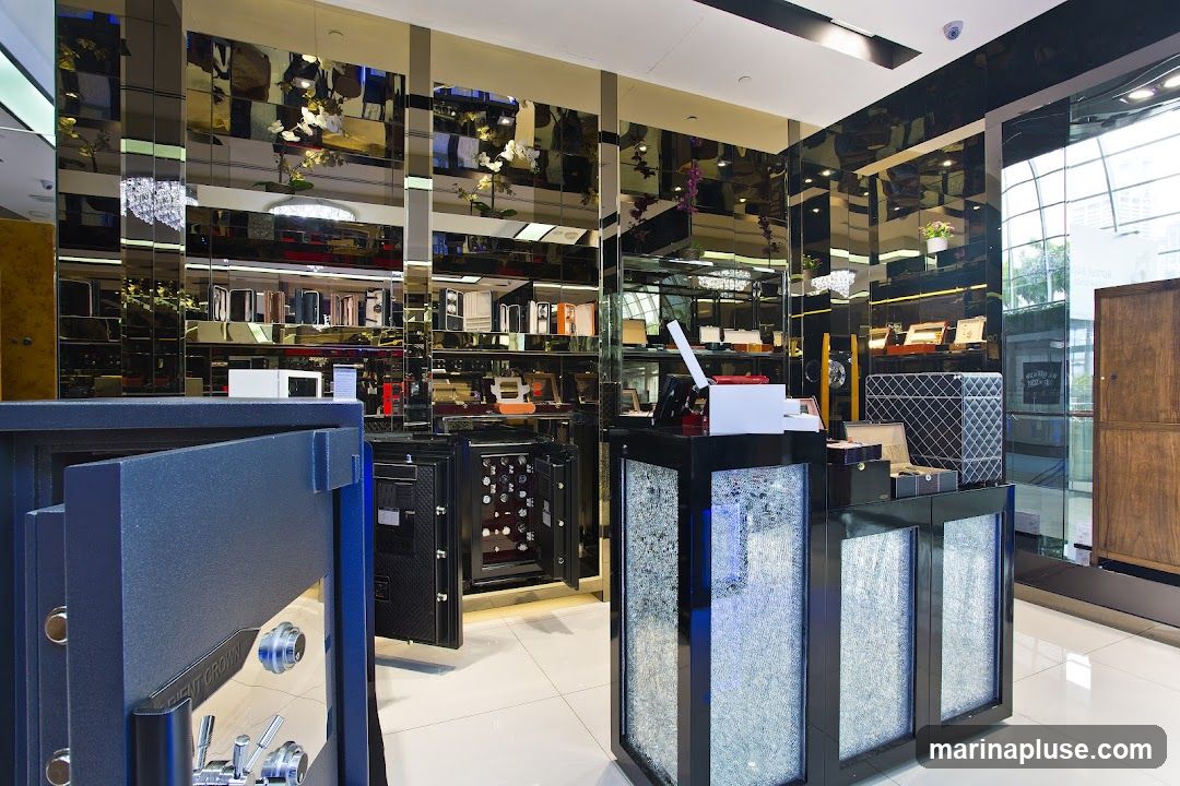 A high-end showroom displaying various safes and luxury storage solutions. In the foreground, a large, dark blue safe is open, revealing its intricate locking mechanism. Behind it, another open safe is filled with watches. The walls are lined with mirrored panels reflecting shelves stocked with boxes and display cases. A central counter features a white point-of-sale system and decorative elements. A tall wooden cabinet stands to the right, and large windows offer a glimpse of the outside world.