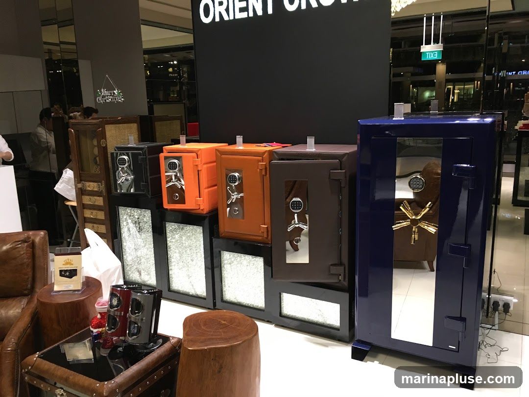 A display of various safes in different colors and sizes, including black, orange, brown, and blue. The safes have digital keypads and metallic accents. In the foreground, there are leather chairs, wooden stools, and a glass-top table with decorative items. A large black sign with white text, partially visible, reads "ORIENT CROWN".