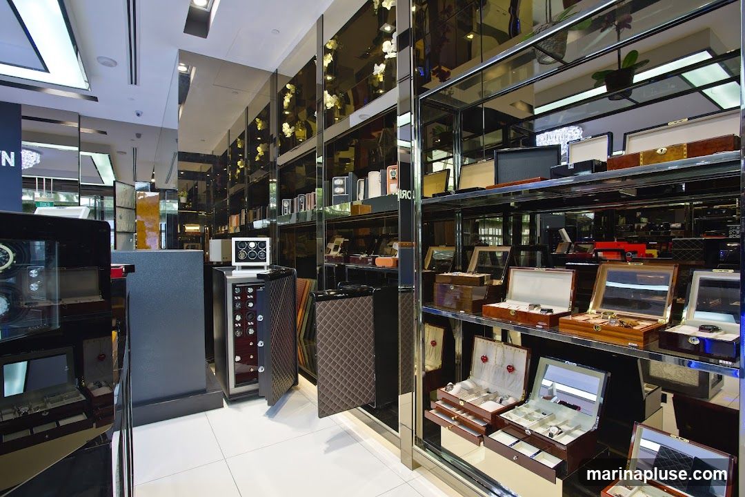 A well-lit interior of a luxury store, showcasing an array of high-end watches and jewelry. The display features polished wooden boxes with glass tops, velvet-lined trays, and intricate watch winders. Mirrored walls and shelves reflect the merchandise, creating a sense of opulence and depth. The overall aesthetic is one of sophistication and exclusivity, with a focus on the detailed craftsmanship of the displayed items.