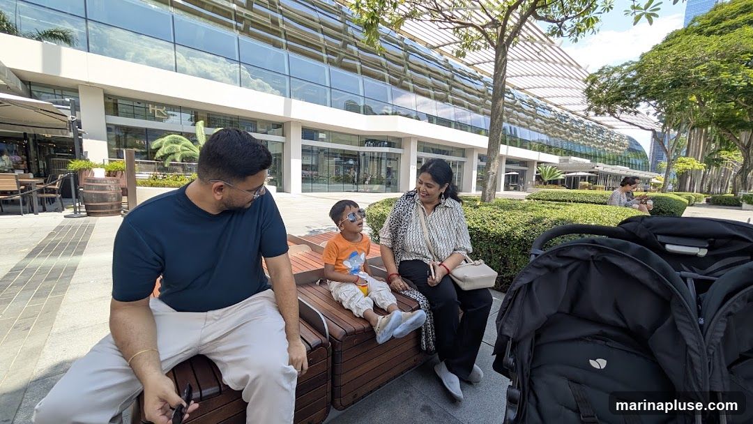 A family, including a man, a woman, and a young boy, are sitting on a wooden bench outdoors. The boy is wearing sunglasses and an orange t-shirt. Behind them is a large, modern building with a glass facade and a distinctive curved roof structure. There are trees and manicured bushes in the foreground and background. Two strollers are visible in the lower right corner.