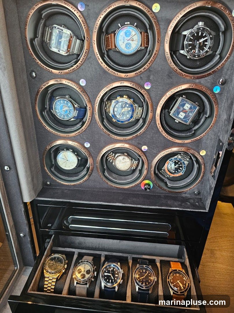 A watch winder displaying nine watches in individual rotating compartments, with an additional five watches displayed in a pull-out drawer below. The watch winder has a dark grey textured finish with copper-toned accents around each compartment, featuring the "ORIENT CROWN" logo. The watches vary in style, including metal bracelets, leather straps, and different dial colors and designs. The drawer below contains watches with metal and leather straps.