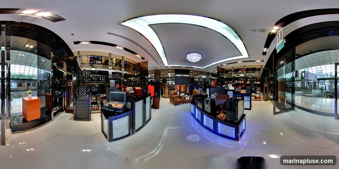 A panoramic view of the interior of a high-end watch store. The store features polished floors, mirrored walls, and a modern ceiling with integrated lighting. Display cases showcase a variety of watches and accessories. In the center, a sign reads "ORIENT CROWN". Comfortable leather armchairs are placed in a seating area. The store appears to be located within a modern shopping mall or airport terminal, with large glass windows offering views of the outside.