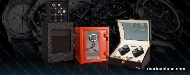 A collection of watch winders and a small orange safe are displayed against a backdrop of large watch faces. The watch winders are in a black unit with multiple compartments and a dark patterned base, and a wooden box with a mirrored interior. The orange safe has a digital keypad and a chrome handle. The background features out-of-focus images of watches with prominent branding.