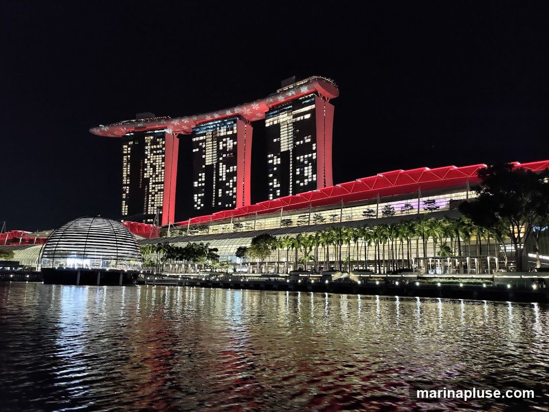 A nighttime view of the Marina Bay Sands complex in Singapore, with its iconic three towers and the convention center. The buildings are illuminated with red and white lights, and the water in the foreground reflects the lights.