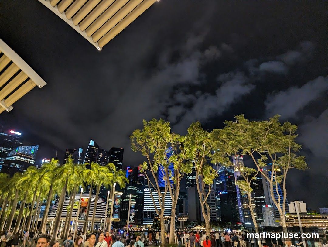A nighttime view of a bustling cityscape with numerous tall buildings illuminated against a cloudy sky. In the foreground, a line of palm trees and other lush greenery are visible, with a crowd of people walking along a promenade. The scene is lit by the city lights and some overhead structures, suggesting an outdoor public space or entertainment area.
