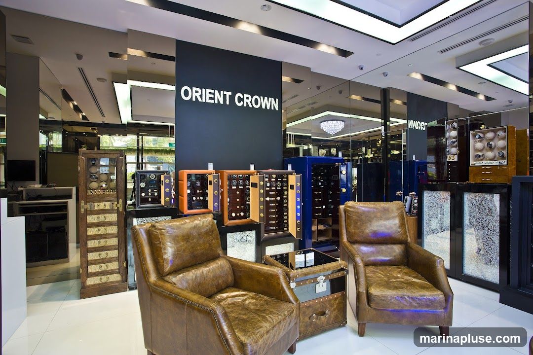The interior of a luxury watch store, featuring the brand name "ORIENT CROWN" displayed prominently on a dark wall. The store is furnished with plush leather armchairs and display cabinets filled with watches. Several watch winders in various colors (orange, brown, blue) are showcased, along with tall wooden display cabinets and modern glass display cases. The ceiling has modern lighting, and mirrored walls reflect the store's opulent ambiance.