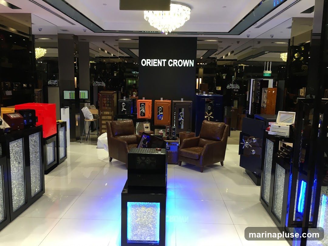 Interior of a high-end showroom displaying various safes and watch winders. The brand name "ORIENT CROWN" is prominently displayed on a black wall. The showroom features plush leather armchairs, illuminated display cases, and a clean, modern aesthetic with reflective surfaces and bright lighting.