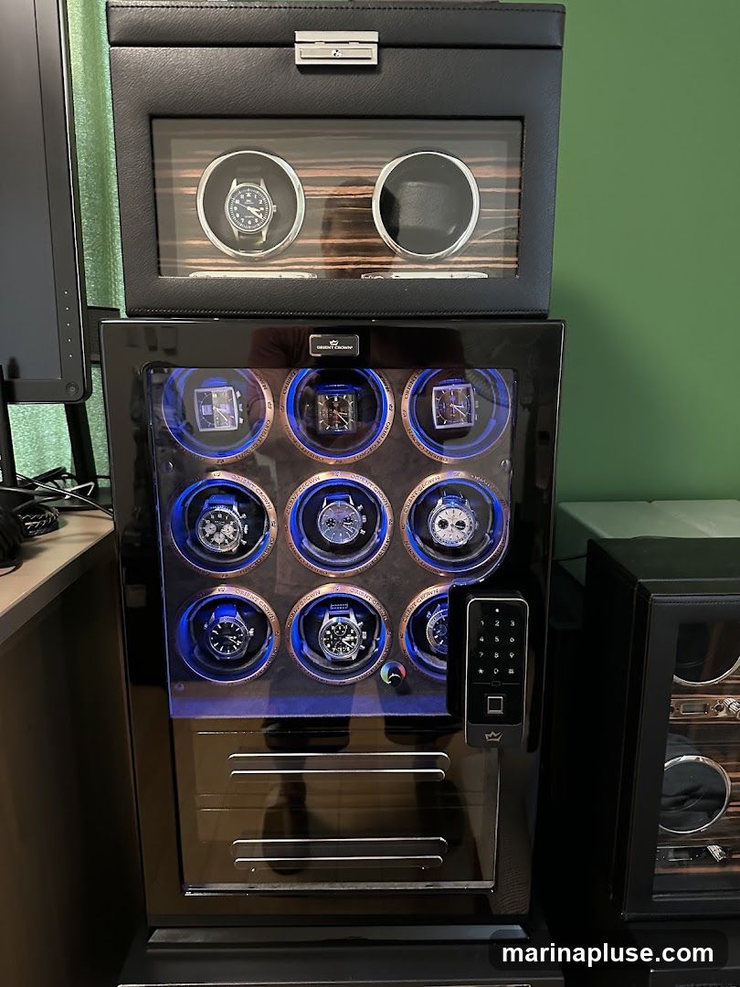 A black watch winder with multiple compartments for watches. The top compartment has a glass lid revealing two watch slots, one with a watch. The main compartment below has nine illuminated watch slots, each displaying a watch. The lower section has a series of horizontal lines, possibly drawers. A keypad and fingerprint scanner are visible on the right side of the main compartment. Another watch winder is partially visible to the right.