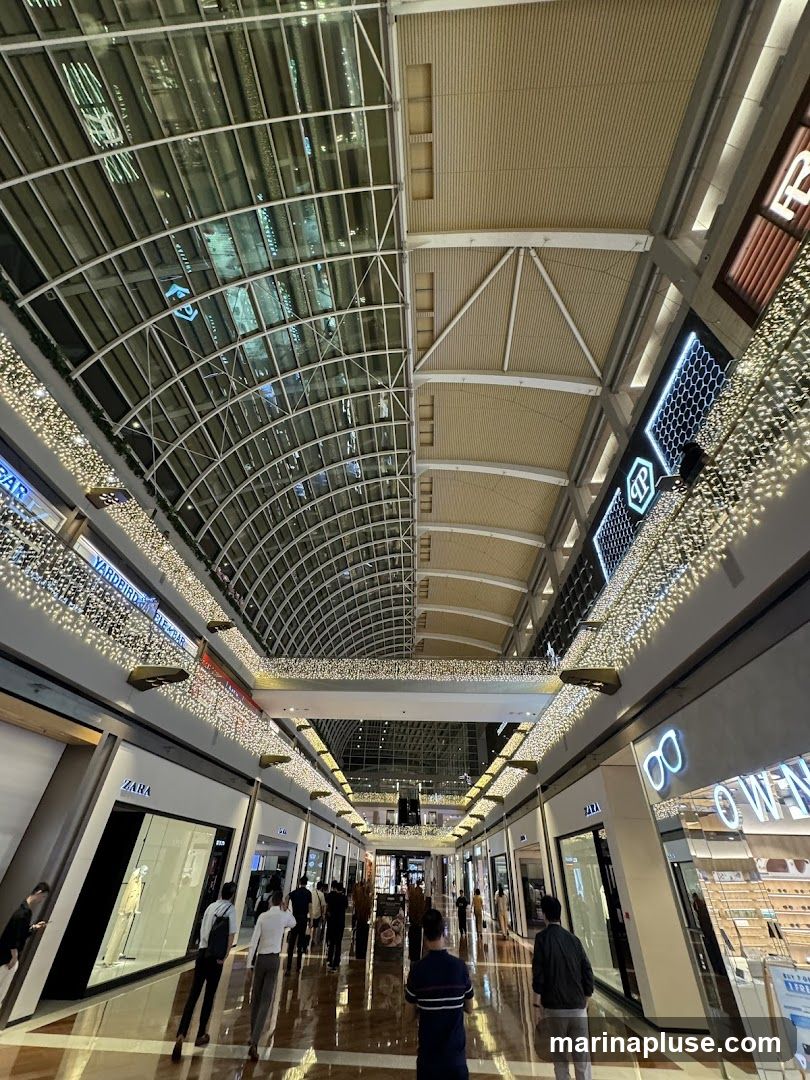A wide-angle view of a modern shopping mall interior with a curved glass ceiling and festive string lights. Shops like Zara and others are visible along the walkway, with shoppers browsing. The architecture features a mix of sleek modern design and warm lighting.