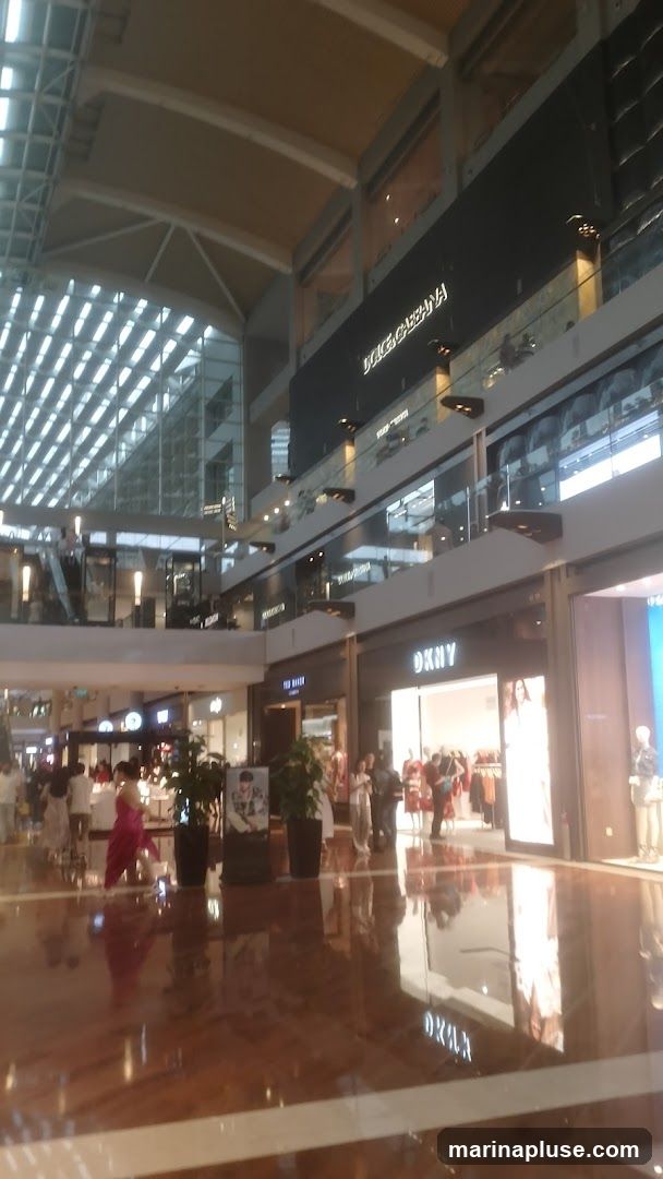 The image shows the interior of a multi-level shopping mall with a high, vaulted ceiling and a glass roof. Several stores are visible on different levels, including Dolce & Gabbana and DKNY. People are walking around the polished floor, and mannequins are displayed in store windows.