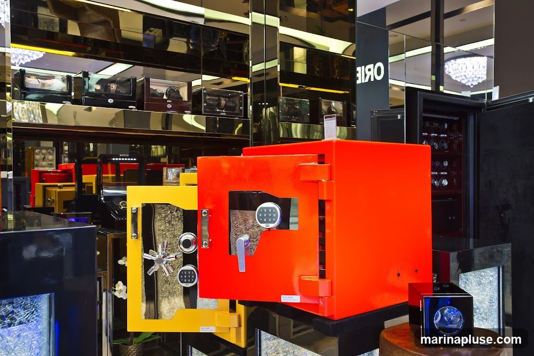A retail display featuring several safes in bright colors, including orange and yellow, with their doors open to reveal internal mechanisms and digital keypads. In the background, shelves are stocked with watch winders and luxury watches. A black safe with multiple watch winders is also visible. The store interior has mirrored walls and a chandelier, suggesting a high-end establishment.