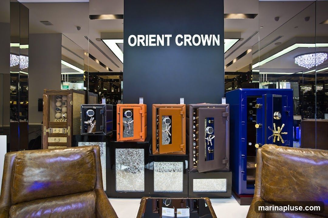 A display of various safes and watch winders inside a store. The brand name "ORIENT CROWN" is prominently displayed on a dark blue wall. In the foreground, two plush brown leather armchairs are visible. The safes are in different colors including black, orange, brown, and blue, with some open to reveal their interiors. A wooden cabinet with multiple watch winders is also part of the display. The background features mirrored walls reflecting the store's interior, including chandeliers and lighting fixtures.