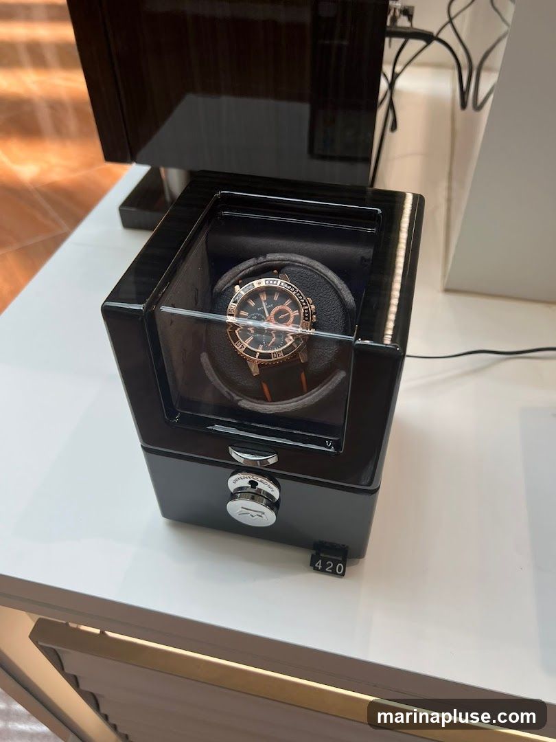 A black watch winder with a rose gold watch inside is displayed on a white surface. The watch has a black dial and a rotating bezel. The winder has a clear lid and a silver knob on the front. A small black tag with the number '420' is attached to the front of the winder.