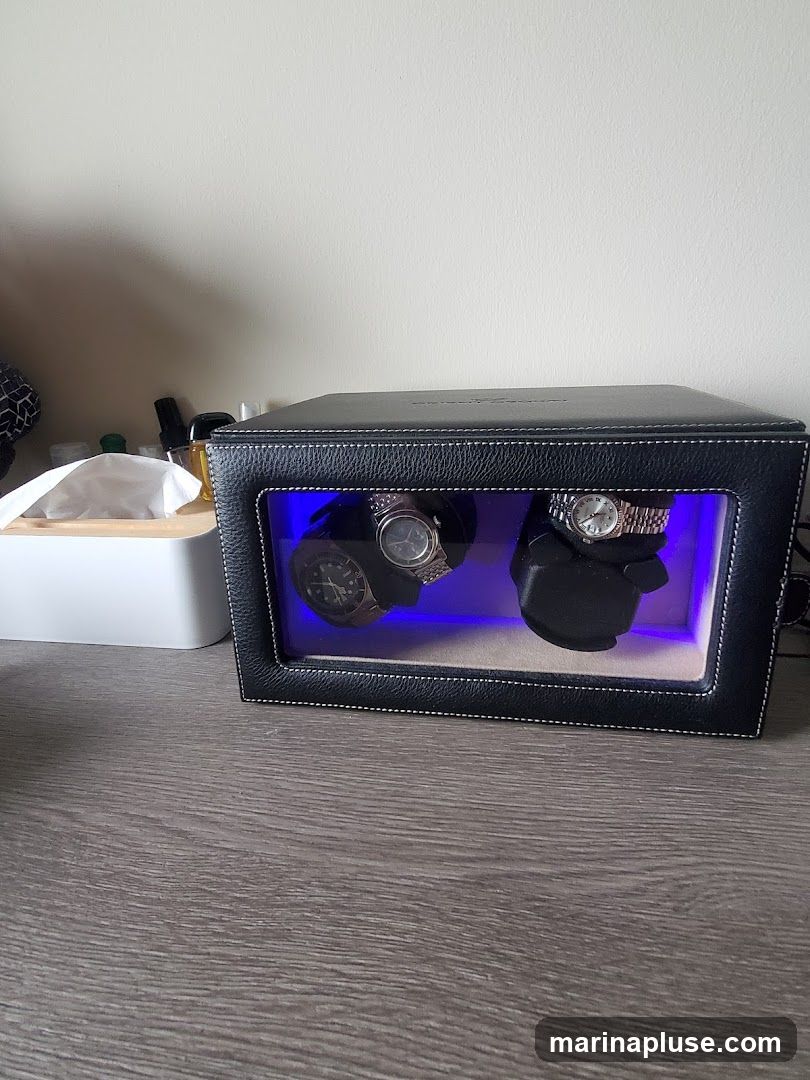 A black leather watch winder with white stitching is shown on a wooden desk. The winder has a glass front, revealing two watches on display, illuminated by blue light. To the left of the winder is a white tissue box.