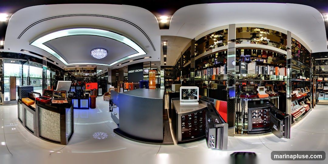 A panoramic view of a high-end store interior, likely selling watches and jewelry. The space features sleek, modern displays with mirrored surfaces reflecting the merchandise. Prominently displayed are various watch cases, jewelry boxes, and what appears to be a safe or display cabinet for luxury watches. The lighting is bright and sophisticated, with a large chandelier visible in the background. The store has a clean, polished aesthetic with a light-colored floor and a dark, curved counter in the foreground. Signage for 'ORIENT CROWN' is visible on a dark panel.