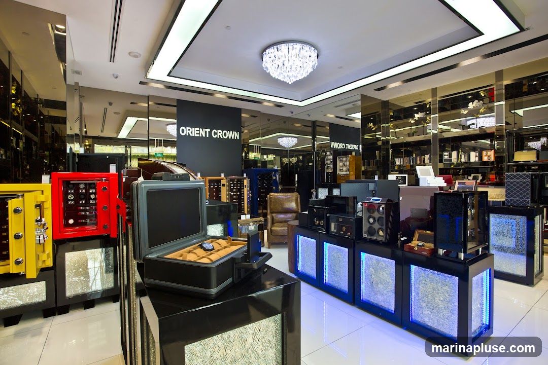 A well-lit interior of a luxury store, possibly a watch or jewelry store, with the name "ORIENT CROWN" visible on a dark banner. The display features several safes in bright colors like yellow and red, a grey case with watches inside, and various watch winders and display cases illuminated with blue LED lights. In the background, shelves are stocked with merchandise, and a plush leather armchair is visible. The overall ambiance suggests a high-end retail environment.