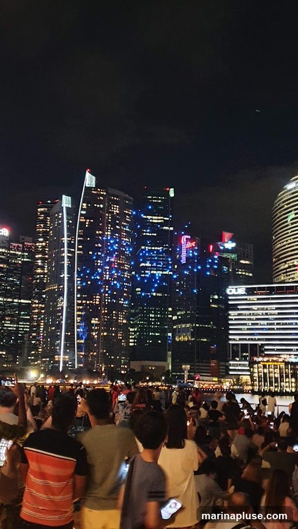 A nighttime cityscape featuring numerous illuminated skyscrapers. Blue lights are visible on some buildings, and a few red lights top others. In the foreground, a large crowd of people are gathered, many holding up their phones, presumably to take pictures or videos of the scene. The sky is dark with a faint airplane trail visible.