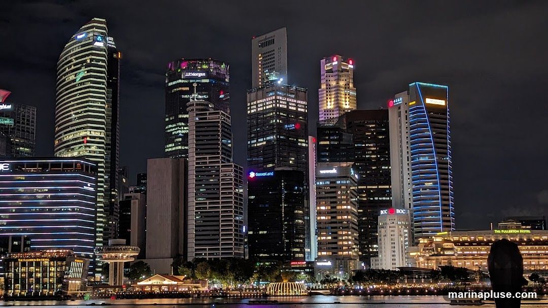 A nighttime view of a modern city skyline with numerous illuminated skyscrapers. The buildings display a variety of architectural styles and lighting, with some featuring prominent company logos such as J.P. Morgan, Guocoland, and Wework. In the foreground, a body of water reflects the city lights, and a low-rise building with the text 'THE FULLERTON HOTEL' is visible.