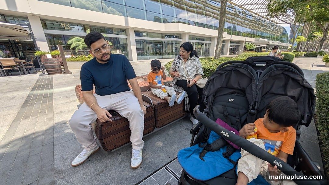 A family is taking a break on a wooden bench outside a contemporary building with large glass windows. A man and a woman are seated, while two young children are also present, one in a stroller. The background shows an outdoor seating area with tables and chairs, suggesting a commercial or dining establishment.