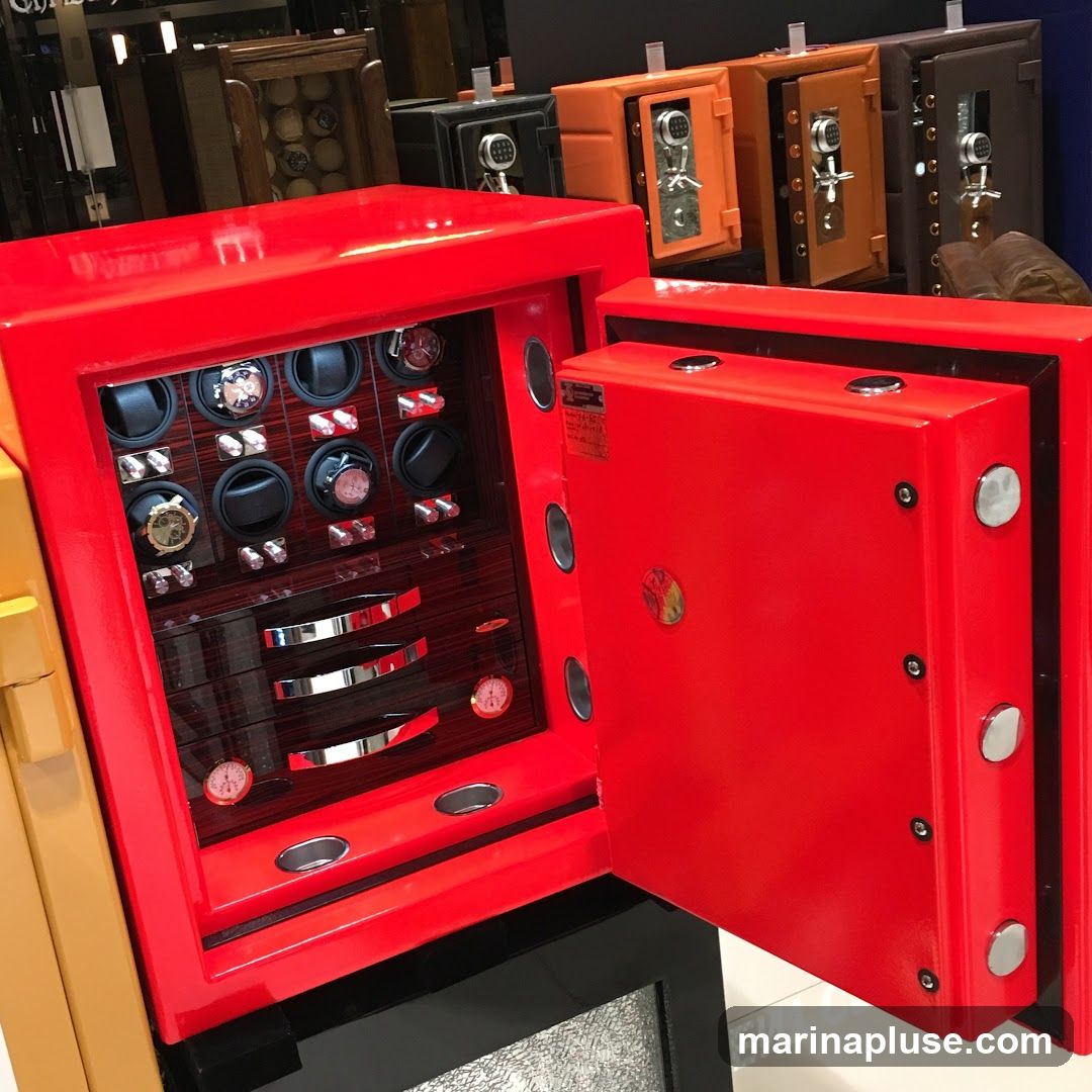 A bright red safe is open, revealing an interior lined with watch winders and drawers. Several watches are visible within the winders. Behind the red safe, other safes in various colors like orange and brown are displayed, along with wooden boxes containing what appear to be billiard balls.